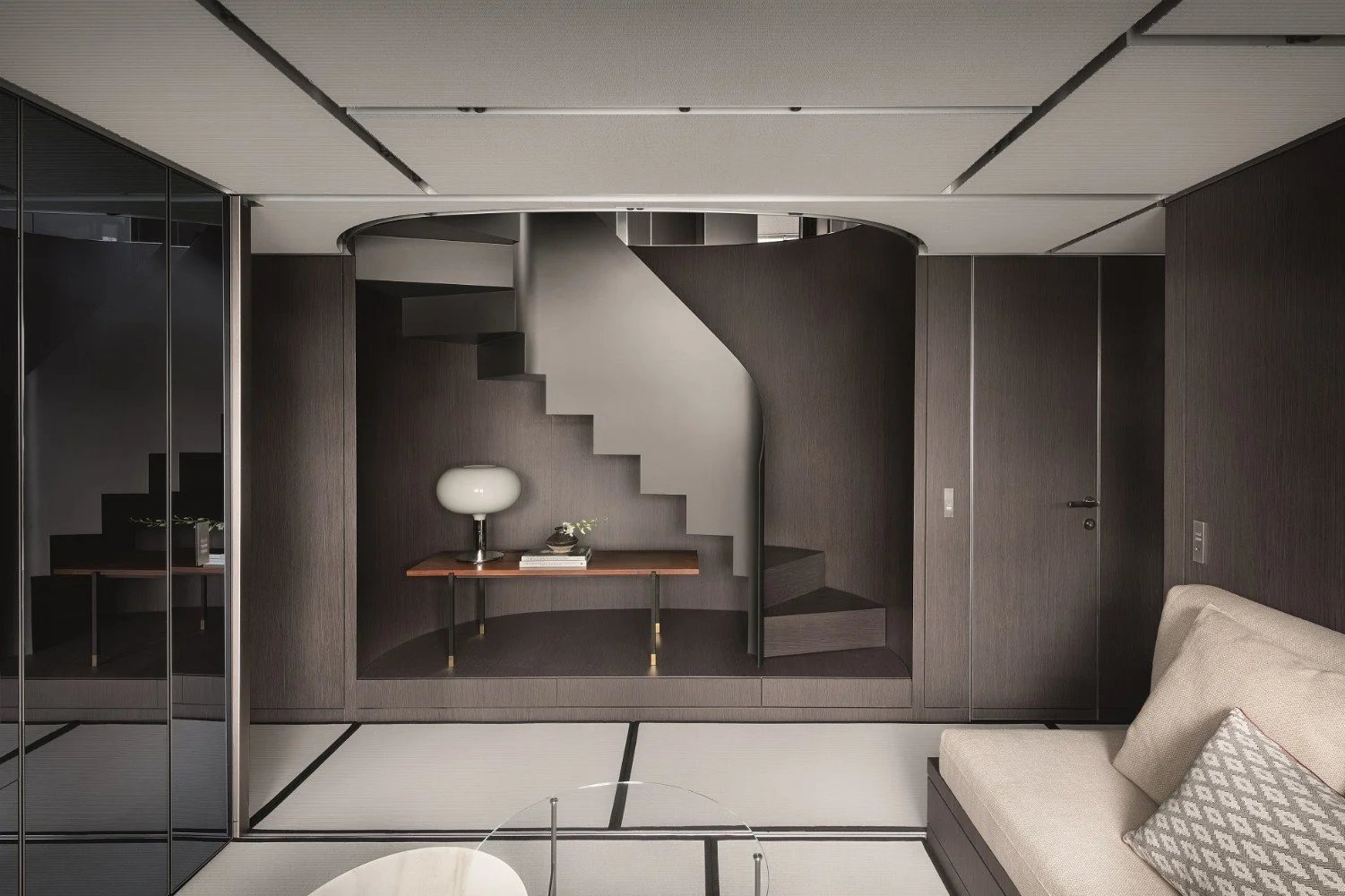 Modern room with a staircase enclosed in a wooden wall alcove, a small wooden table with a white lamp, a few books, and a small plant, a beige sofa with pillows, and mirrored closet doors reflecting part of the room.