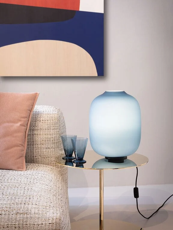 A glass table with decorative items, including a large blue and white lamp, two blue drinking glasses, a beige textured sofa with a pink pillow, and an abstract painting on the wall behind.