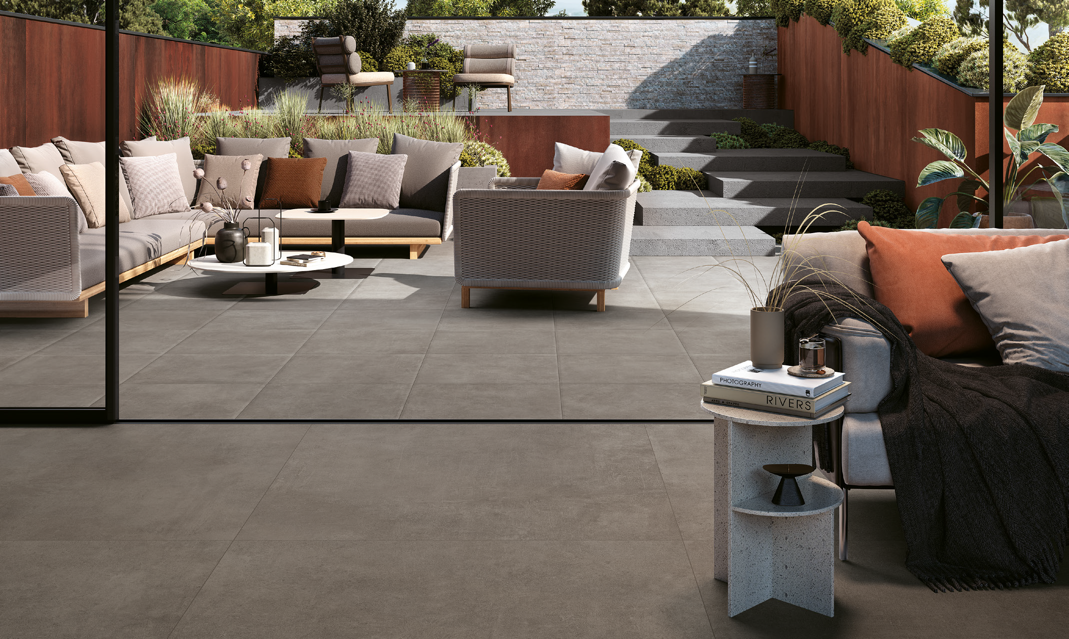 Modern outdoor patio with comfortable seating, including a sofa and armchair, decorated with cushions, with a coffee table holding decorative items. Behind, there are stairs leading to a higher garden area with shrubs and flowers, surrounded by reddi