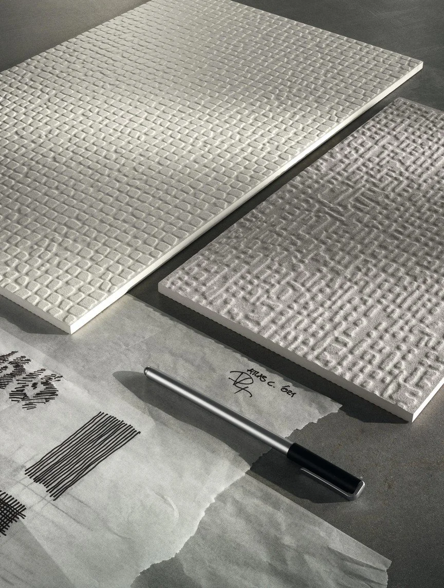 Two textured, rectangular sample tiles or panels, one white and one grey, placed on a grey surface, with a pen and a piece of paper with handwritten notes and barcode sketches nearby.