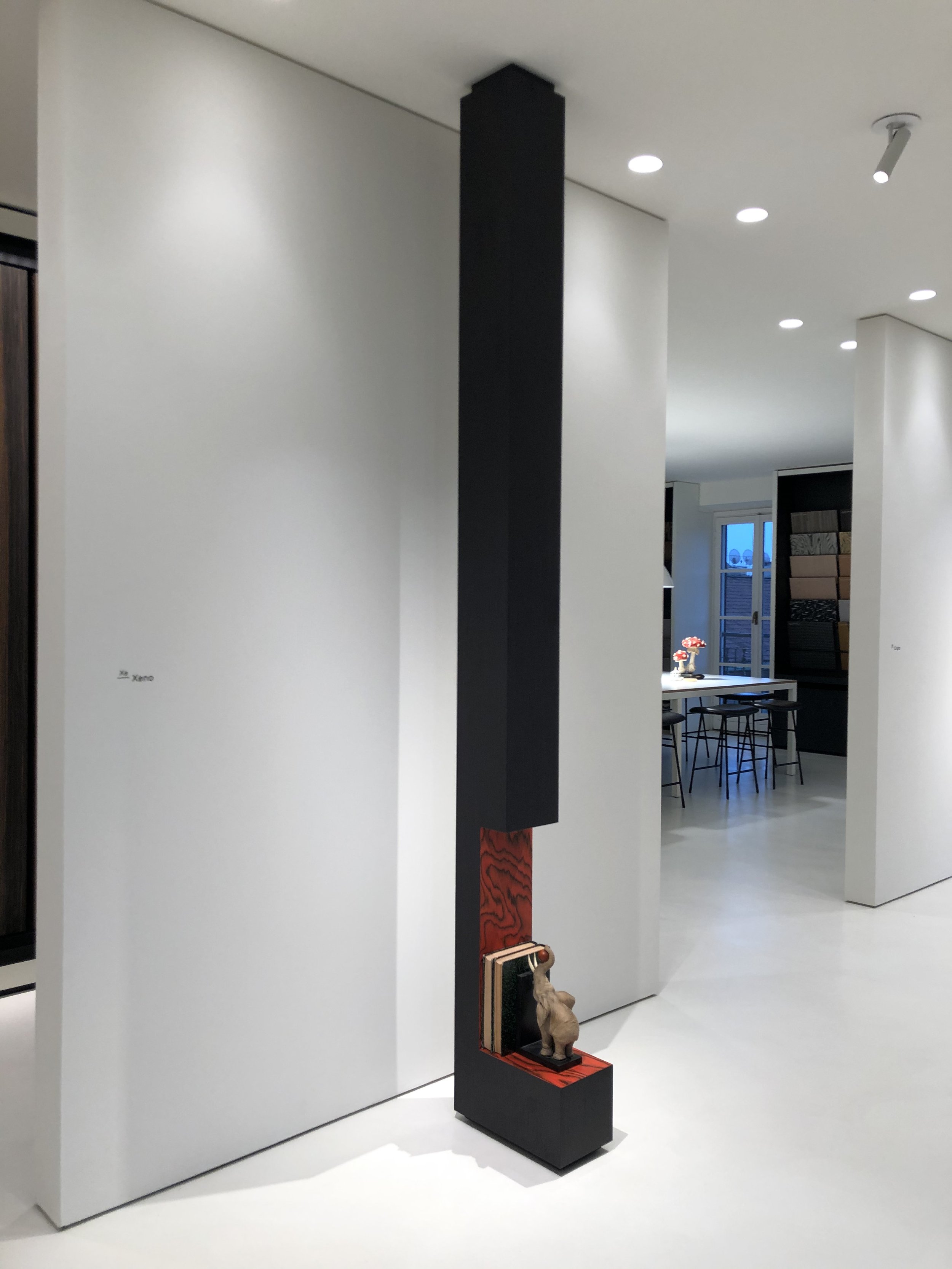 A black vertical sculpture featuring a small animal figure on a red platform at its base, set against white walls in an art gallery.