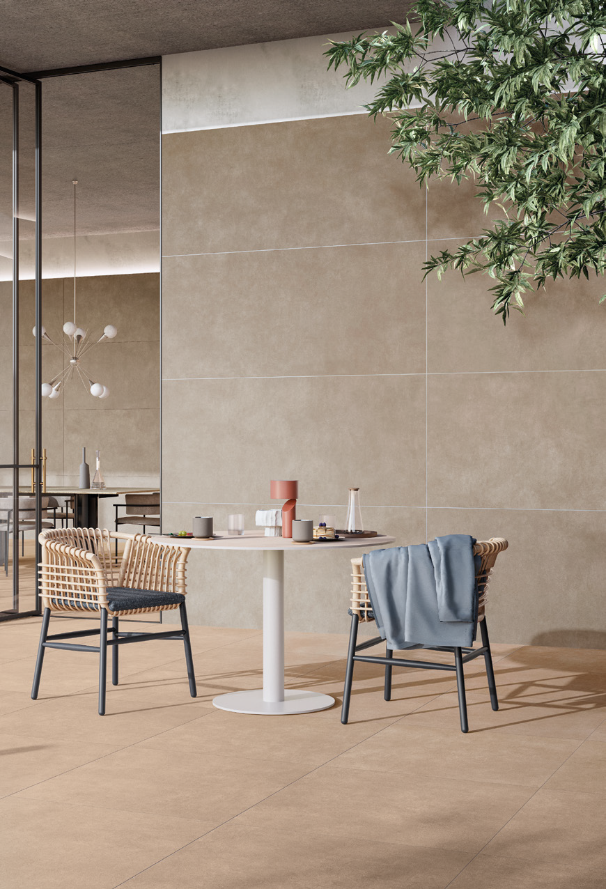 Modern dining area with a round white table surrounded by four rattan-style chairs, one with a blue cloth draped over it. Decor includes a pink lamp, cups, glasses, and a tray. A large green plant is in the upper right corner, with beige wall panels 