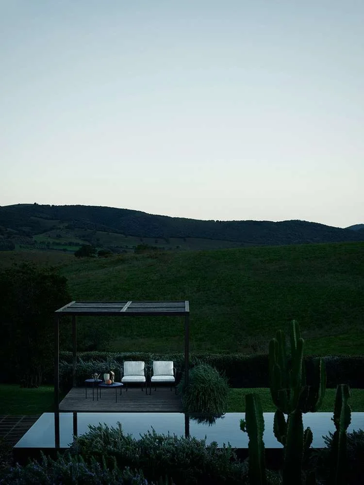 Outdoor seating area with two white chairs and a small table, set on a deck over a reflective pool, surrounded by desert plants, with rolling green hills under a clear sky.