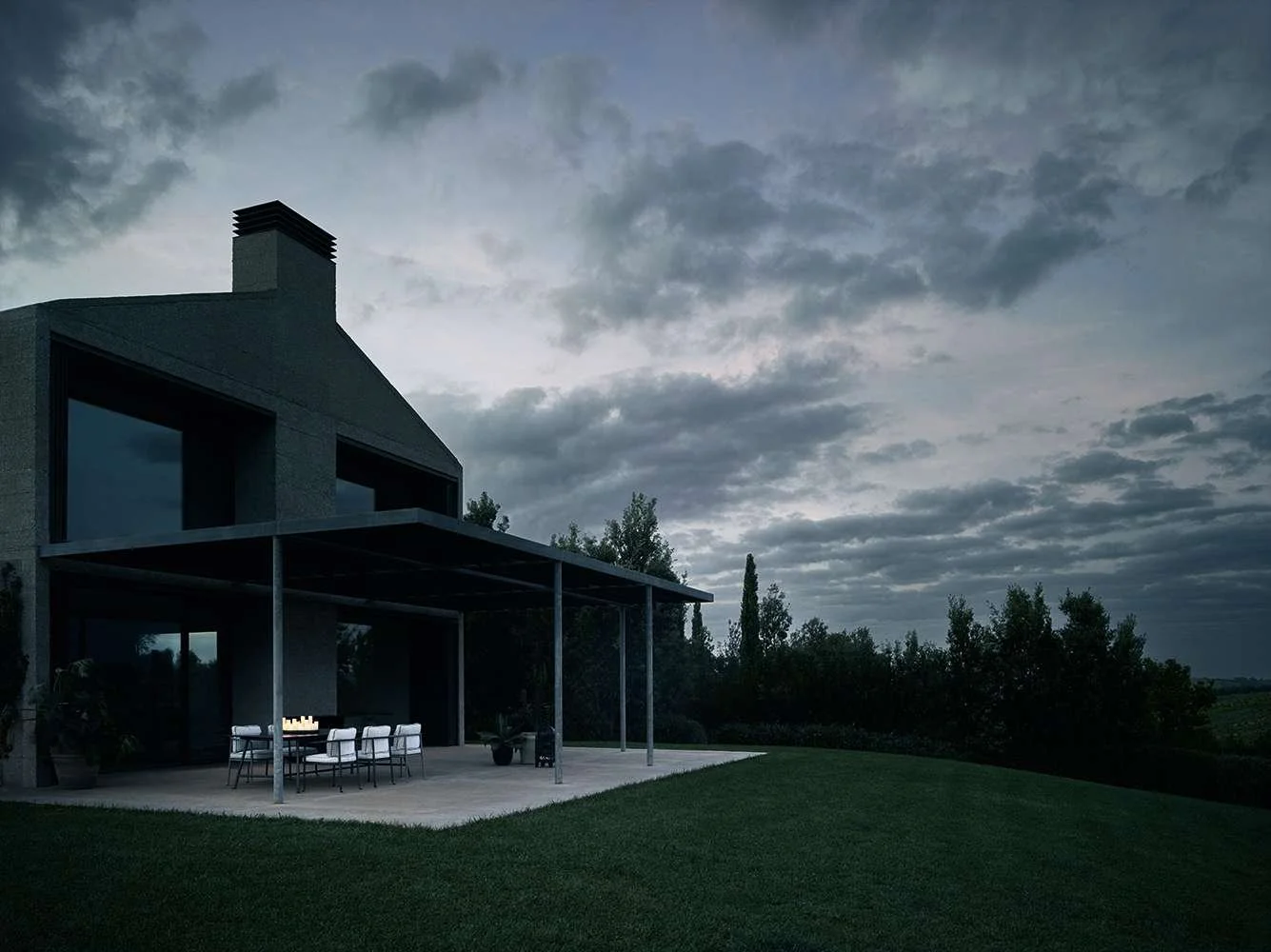 A modern house with a covered patio, dinner table, and chairs, under a cloudy sky at dusk, surrounded by greenery.