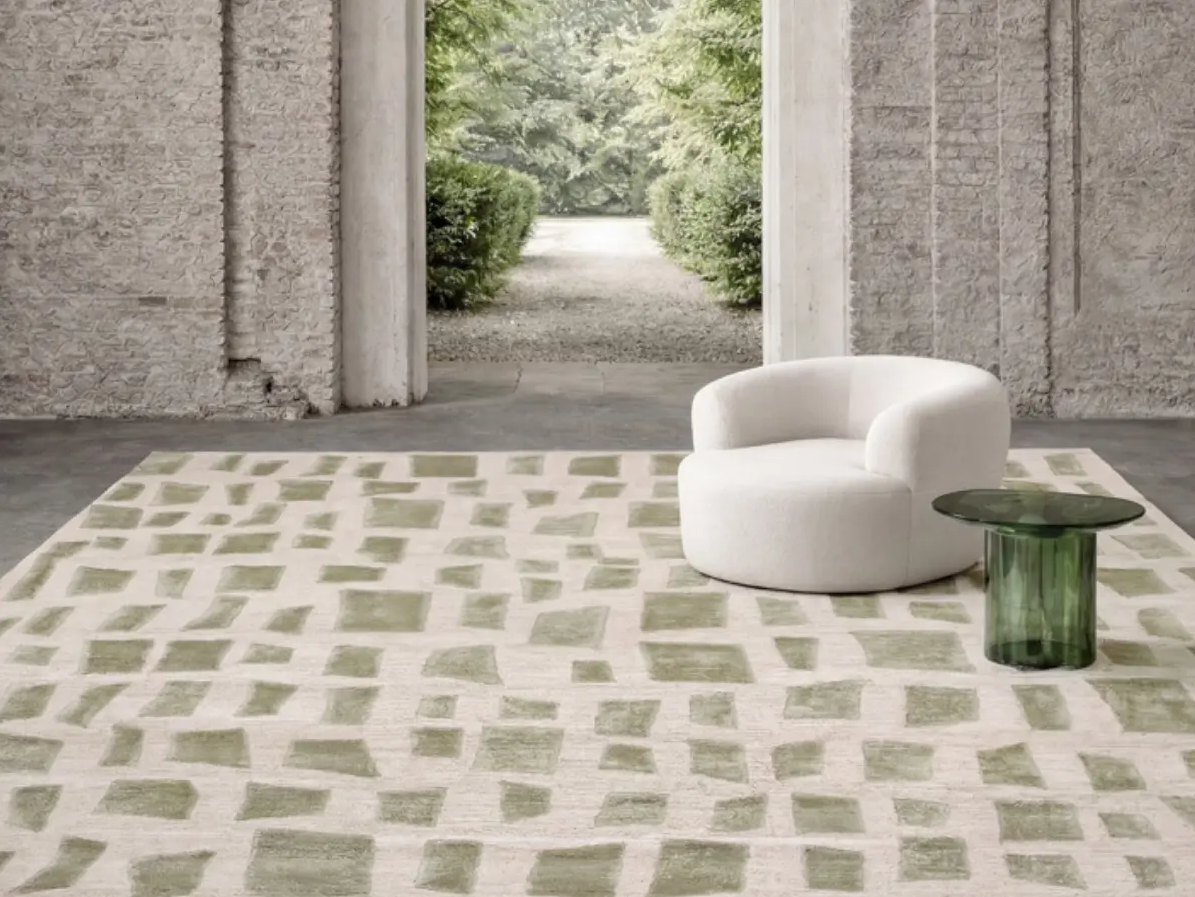 A room with a large window opening to a garden with trees and bushes, a white upholstered armchair, a green glass side table, and a patterned area rug.