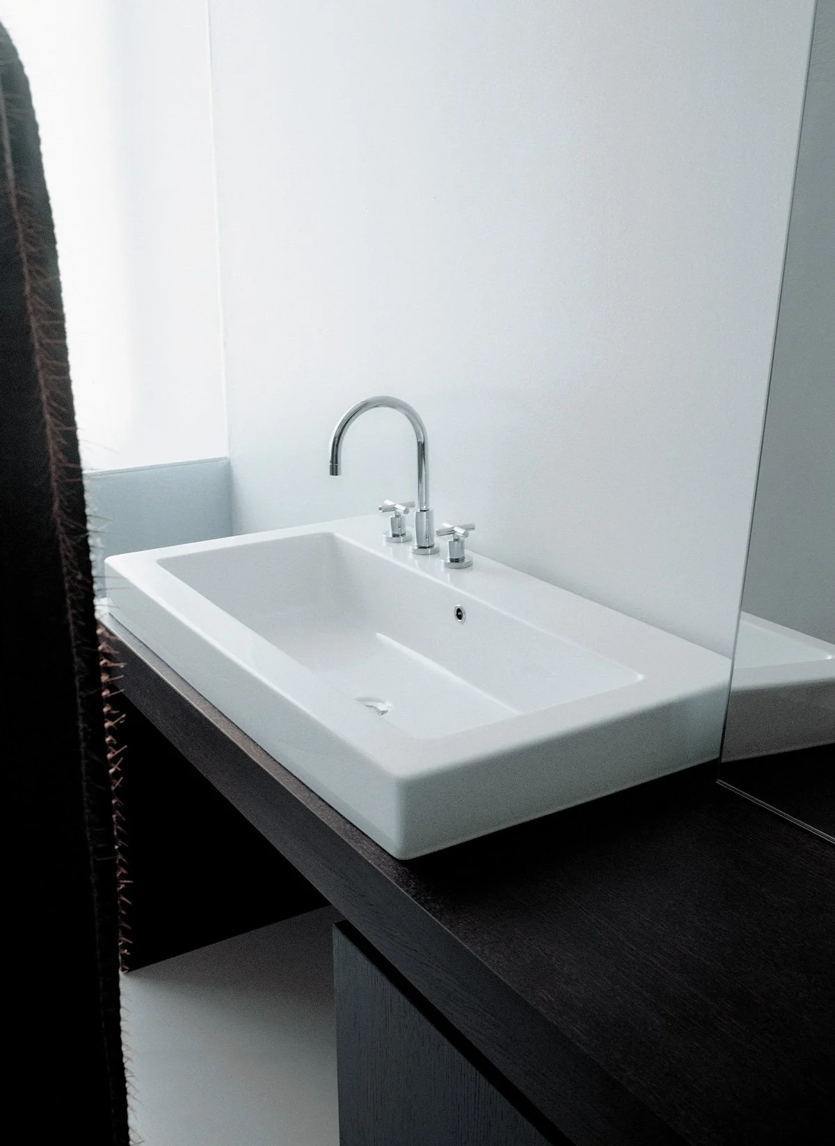 A modern white rectangular bathroom sink with a sleek silver faucet, mounted on a dark wooden vanity, positioned against a plain white wall with a mirror to the right.