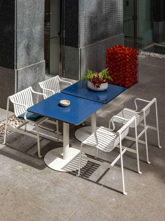 Outdoor patio table with a blue top and four white chairs, adorned with a small plant in a white pot, next to a modern building with gray walls. In the background, a decorative container filled with red balls.