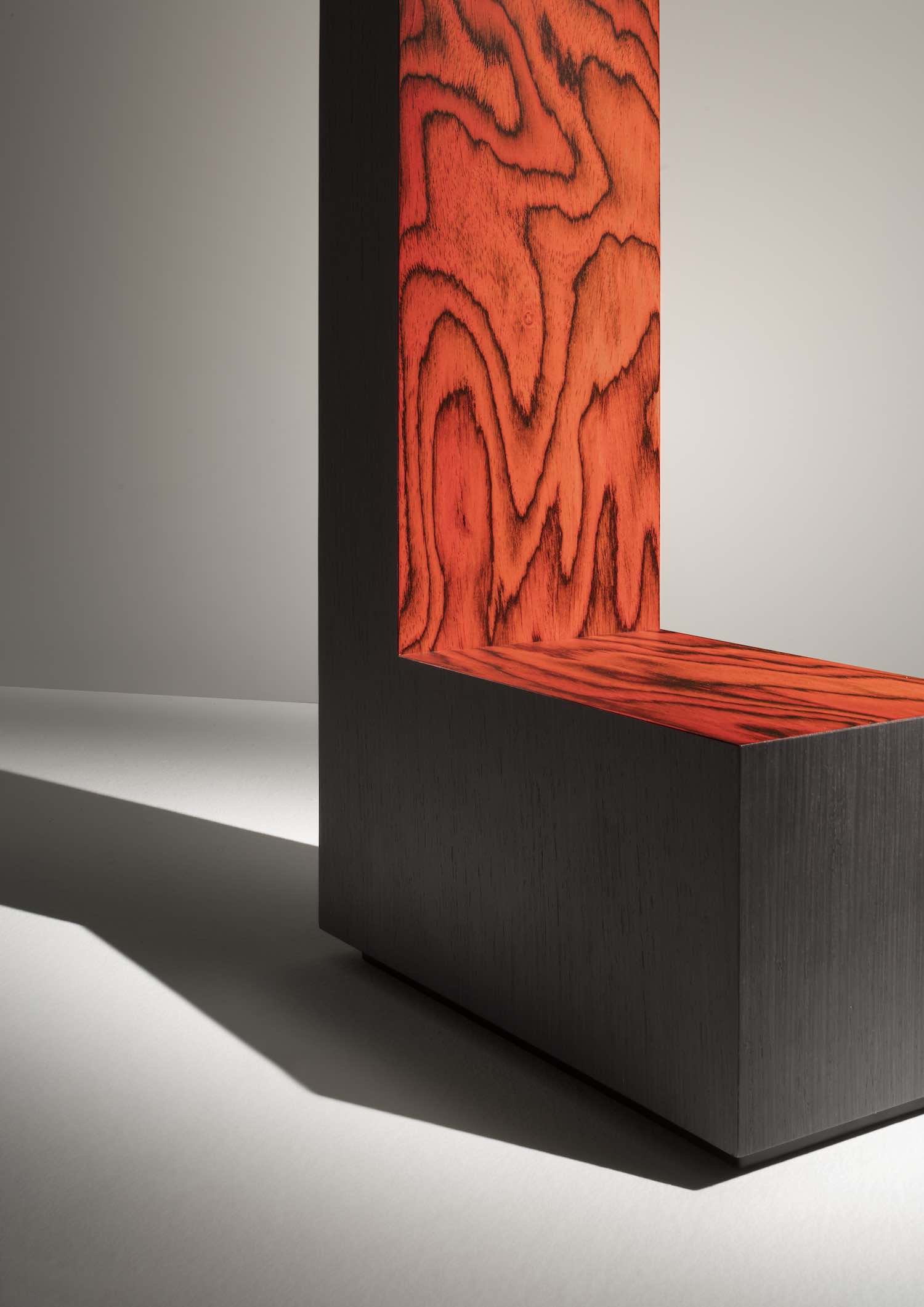 Close-up of a corner of a modern piece of furniture with a dark base and a red panel with black wood grain pattern, casting a shadow on a white surface.