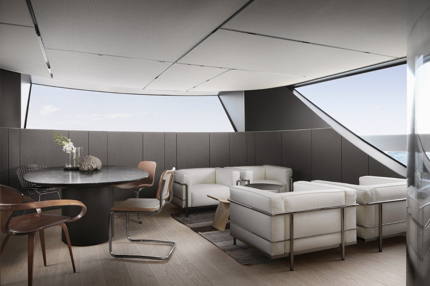Modern interior lounge area with white sofas, a black round table, and a dining table with chairs, large windows showing an ocean view, and a minimalistic decor.