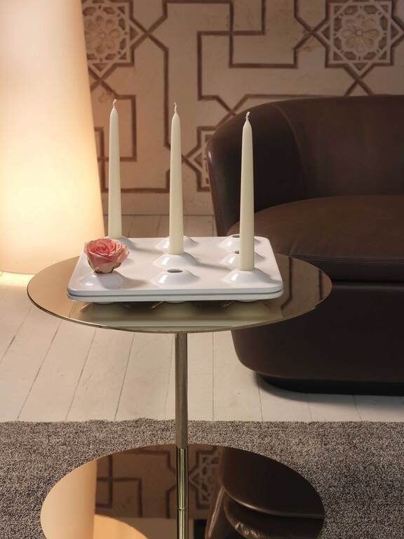 A round side table with three white candles in holders, a pink flower, and a white ceramic tray, situated next to a dark sofa in a living room with patterned wall decor.