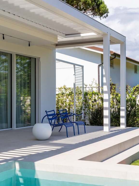 Modern backyard patio with blue chairs, a round white side table, and a swimming pool in the foreground. The patio has a white pergola and a glass sliding door leading into the house. There are green plants and trees in the background.