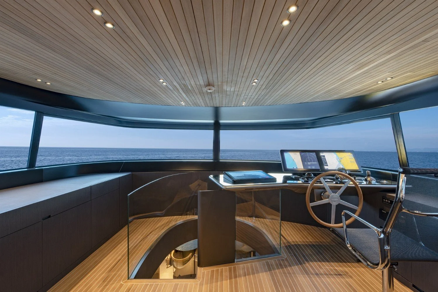 The interior of a yacht's bridge with a panoramic ocean view, featuring wooden ceiling, modern dashboard with screens, steering wheel, and seating area.