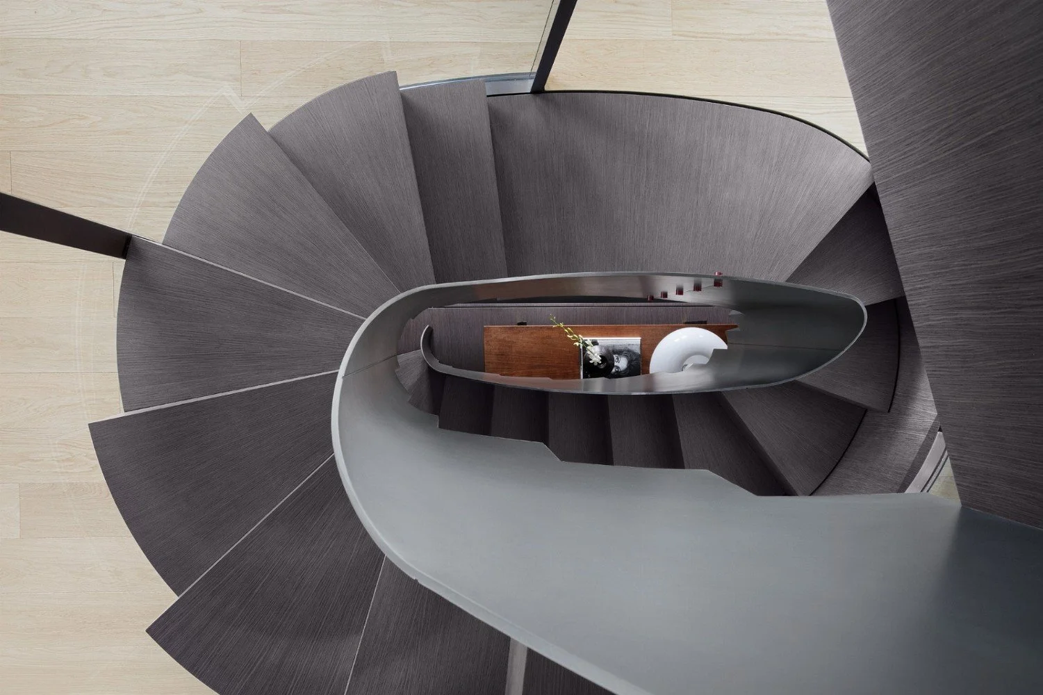 Top-down view of a spiral staircase with dark gray steps and a black handrail, looking downward towards a wooden table with a decorative vase and a white spherical sculpture at the bottom.