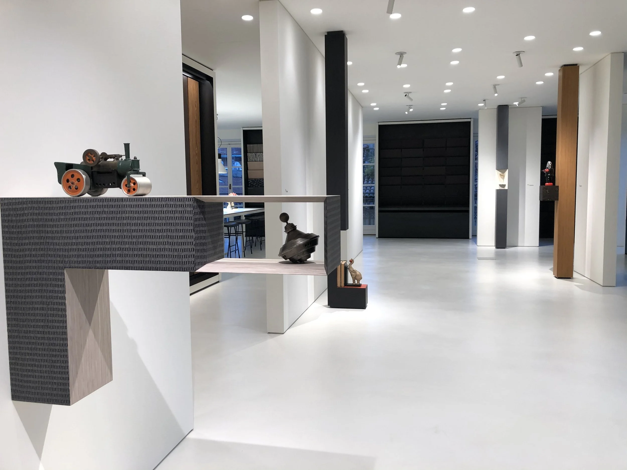 Interior of a modern, minimalist building with white walls, ceiling lights, and decorative art pieces including miniature vehicles and a giraffe sculpture, with large windows in the background.