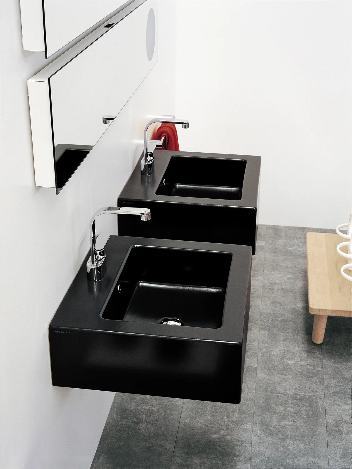 Two black sinks with silver faucets, mounted on a white wall in a clean, modern restroom with gray tiled floor and a small wooden bench with white cup holders on the right side.
