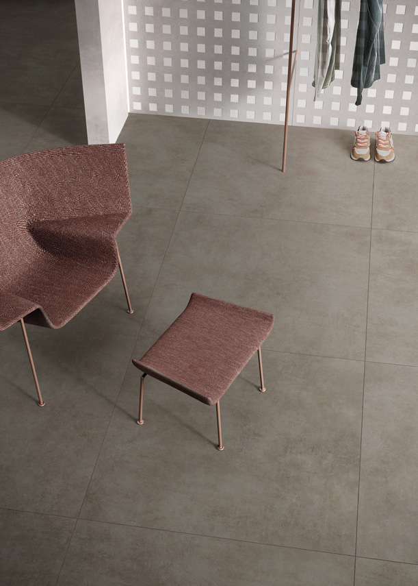 A modern indoor space with large gray tiled floor, a pink upholstered chair, a matching pink ottoman, and a wall with a cut-out pattern. In the corner, there are hanging striped and plaid shirts, and a pair of sneakers on the floor.