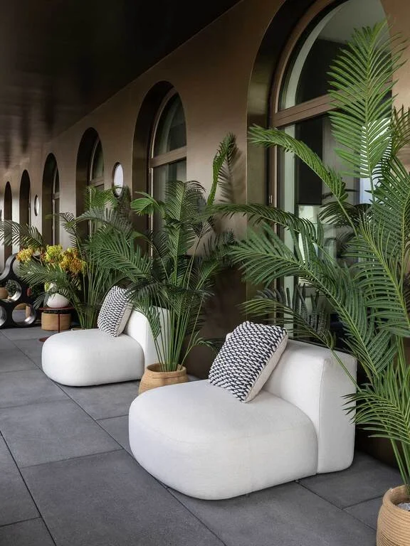 Modern indoor waiting area with white curved chairs, patterned pillows, large potted green plants, and arched windows along the wall.