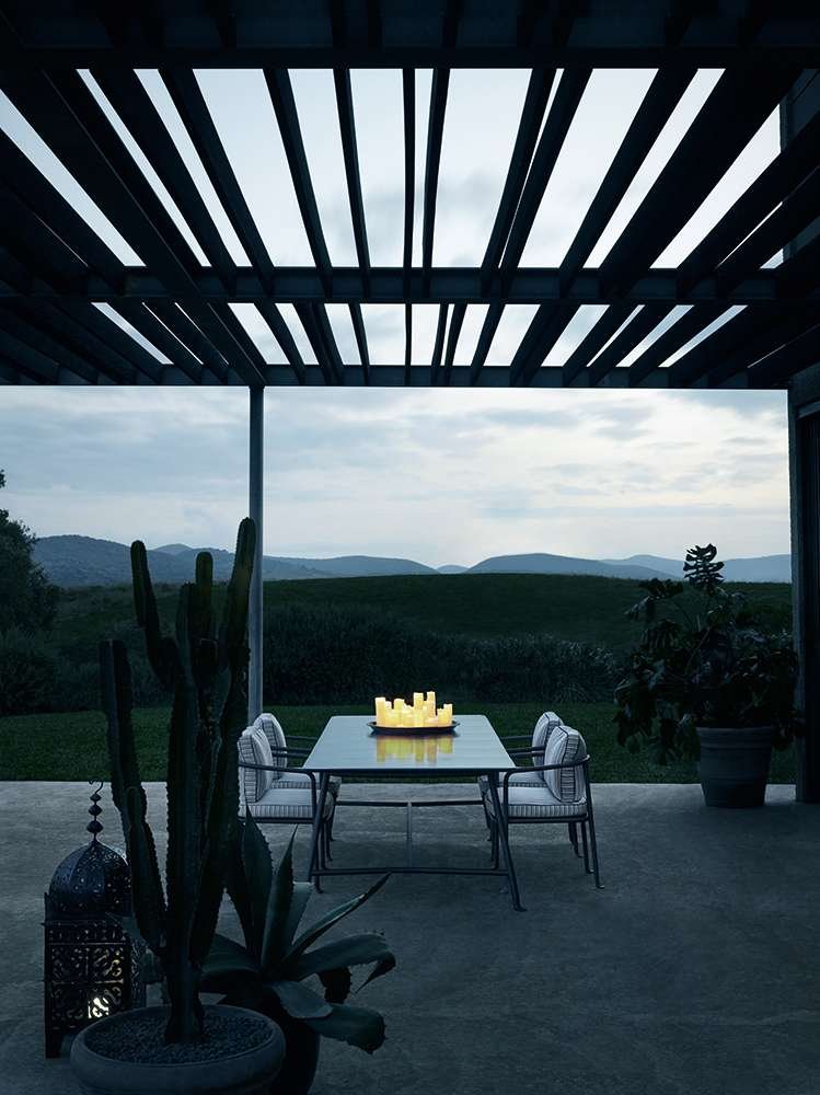 Outdoor patio with a dining table and chairs, candles on the table, potted plants, desert landscape with mountains in the background, and a slatted wooden pergola overhead.