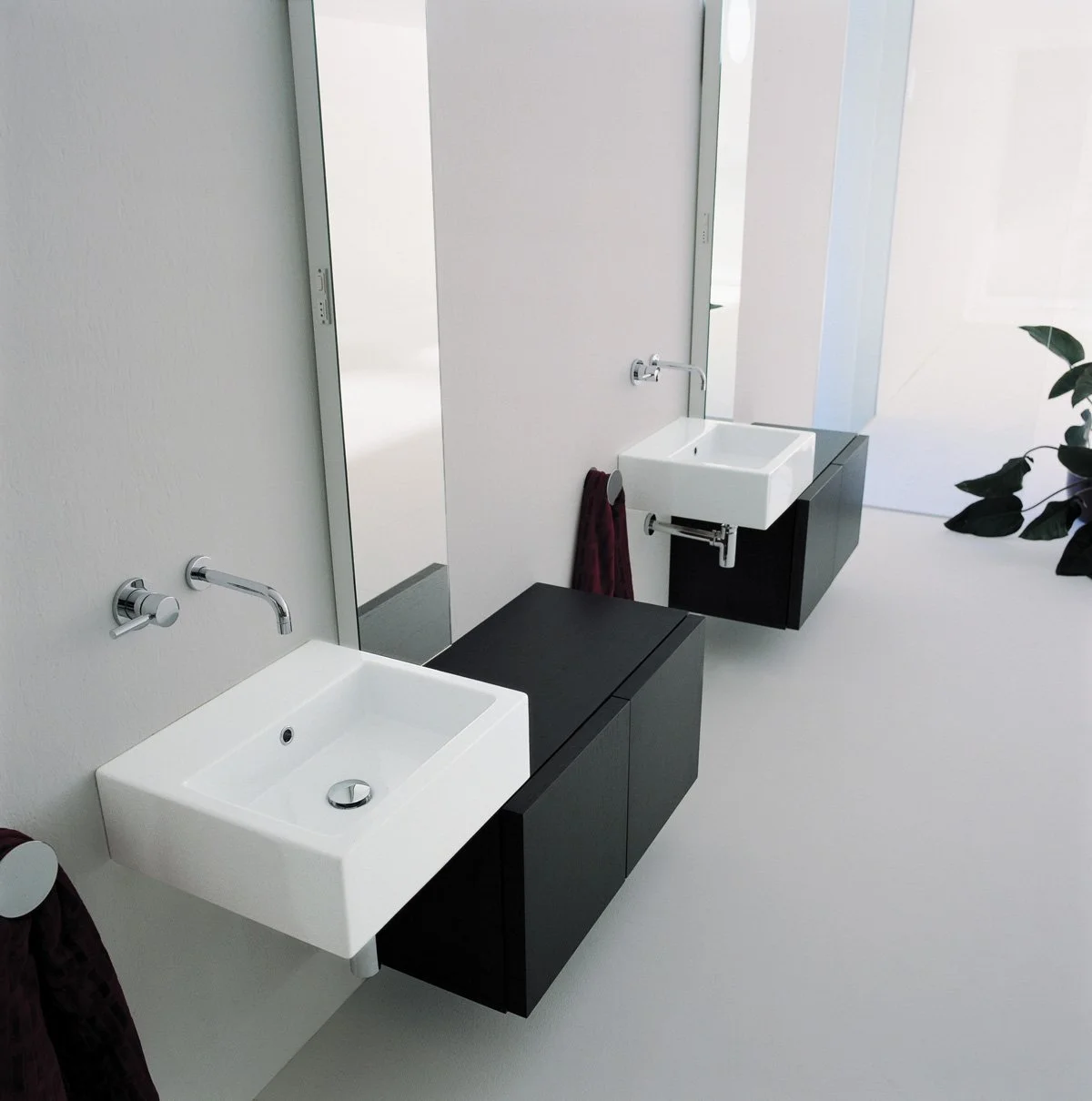 Minimalist bathroom with two black vanities, each with a square white sink, mounted on a white wall, with a large mirror reflecting the sinks, a small towel hanging on a ring, and a potted plant visible in the background.