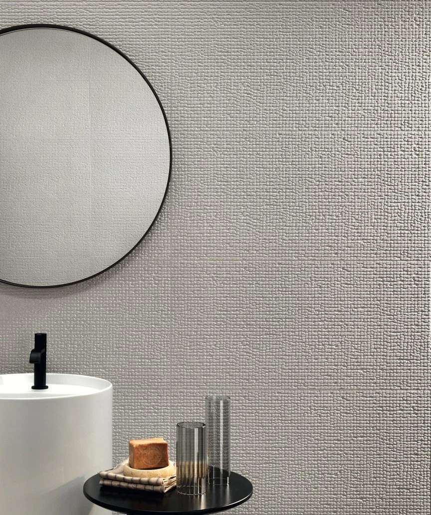 Minimalist bathroom sink area with a white round sink, black faucet, and a black circular side table holding a bar of soap and three empty glass cups, against a textured white wall.