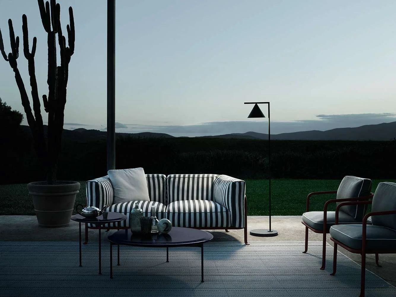 Living room area with a striped sofa, two black chairs with wooden armrests, a tall floor lamp, and a large potted cactus, set against a scenic mountain view at dusk.
