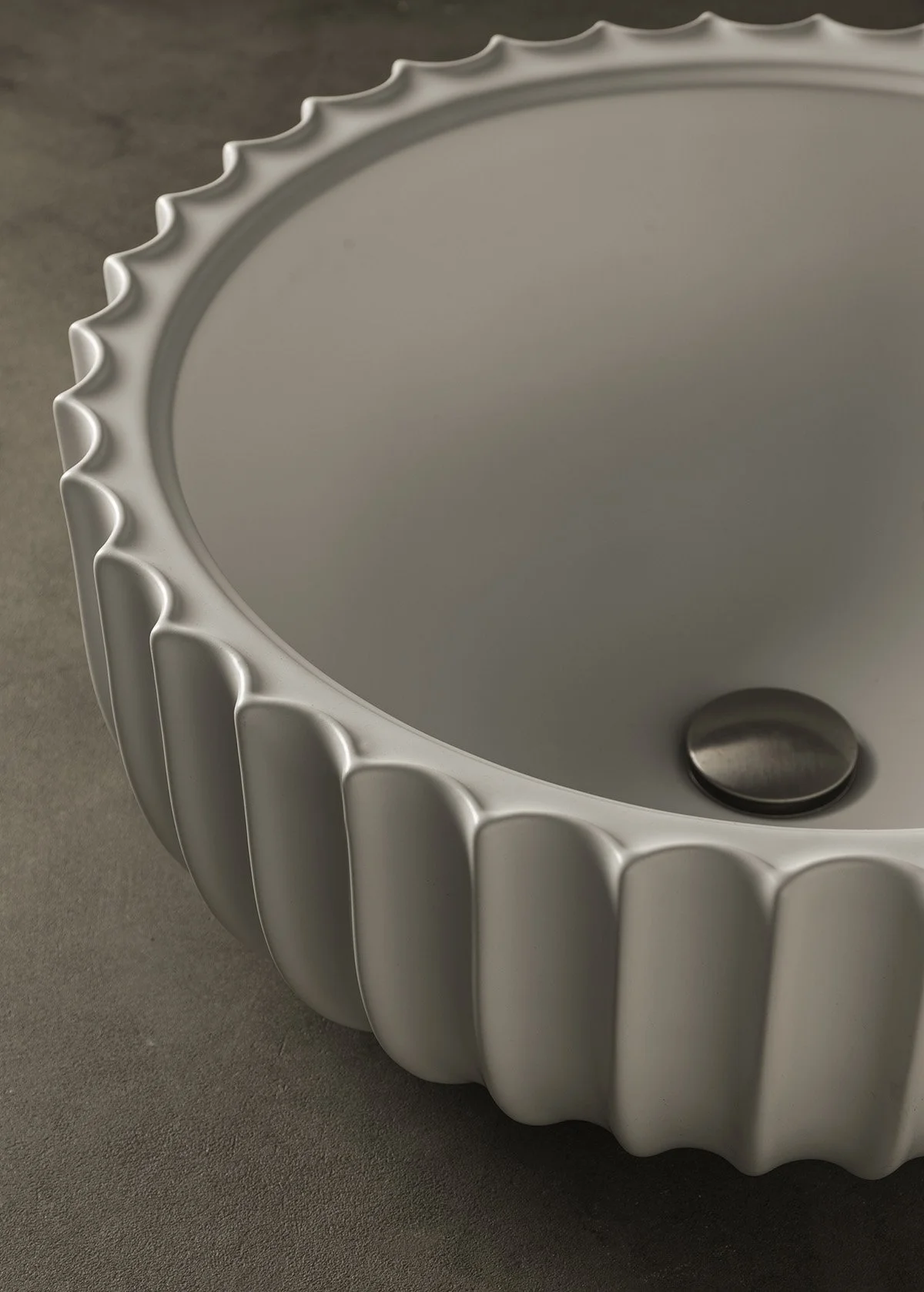 Close-up image of a round, white ceramic sink with a textured, scalloped edge and a silver drain in the center, placed on a gray surface.