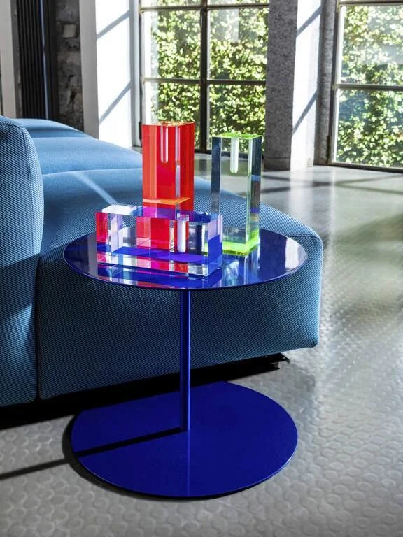 Colorful acrylic blocks on a blue side table near a blue sofa in a bright room with large windows and green foliage outside.