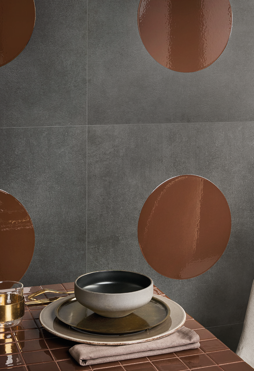 A modern dining table setup with a layered plate, black and white bowl, gold-rimmed glass, and folded napkin, against a wall with large circular brown accents.