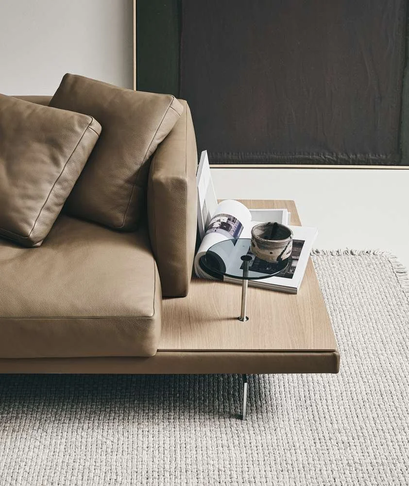 A beige couch with cushions on a wooden sofa table, holding a magazine and a ceramic bowl, next to a black chalkboard or dark wall and a wall with white paint, with a textured cream-colored rug on the floor.