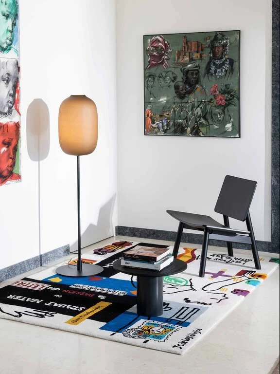 Modern sitting area with a black chair, a black round coffee table with books, a tall tan floor lamp, a colorful abstract rug, and framed artwork on white walls.
