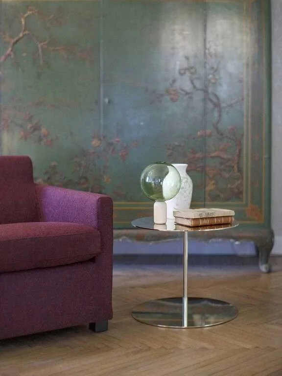 A living room corner with a purple sofa, a small round metallic side table, a textured ceramic vase, a green glass globe, and a stack of books against a decorative painted wall with floral motifs.
