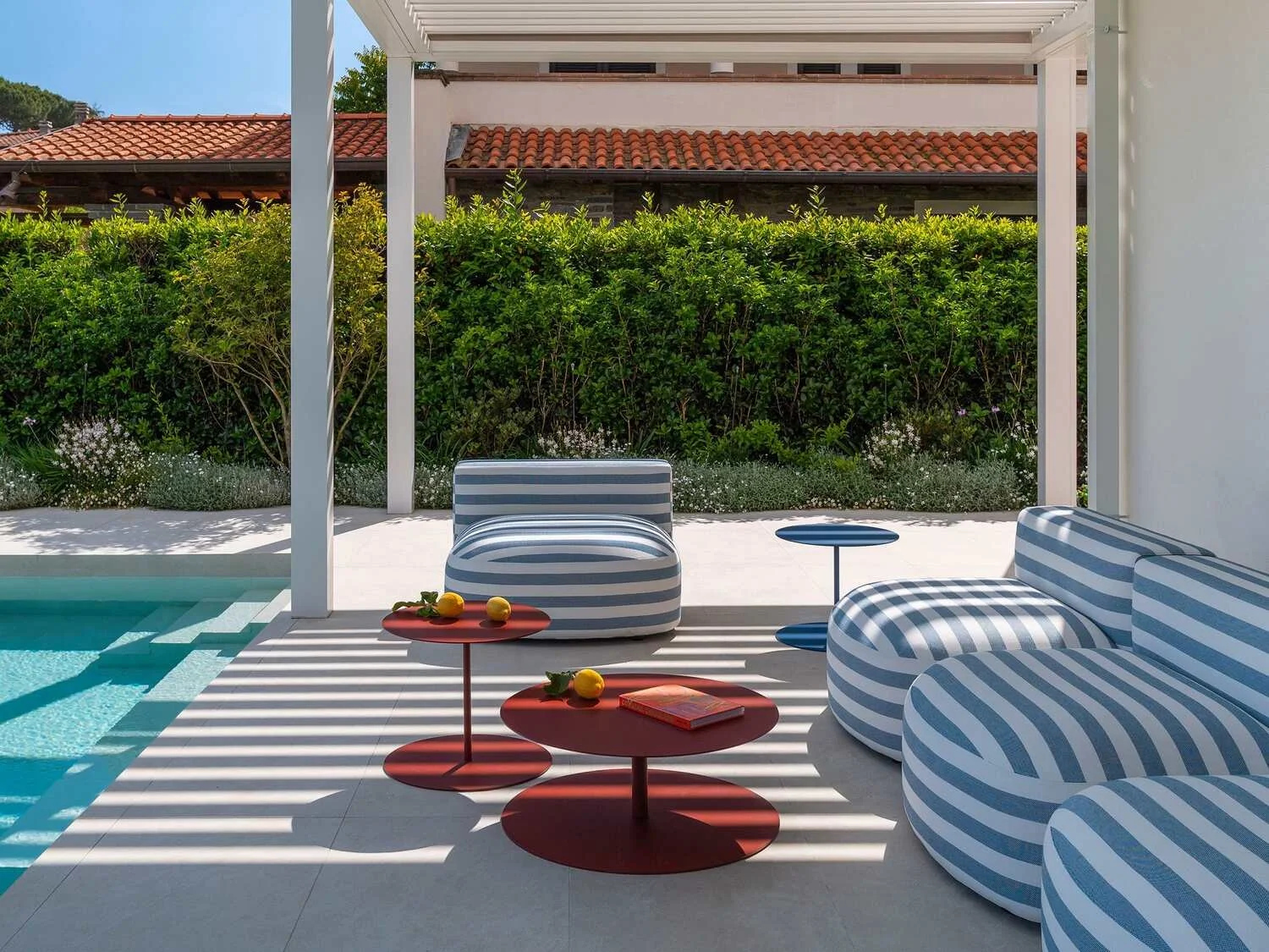Patio with striped blue and white furniture, red side tables with lemons and a book, shaded by a pergola, next to a swimming pool, with lush green bushes and a house with a red-tiled roof in the background.