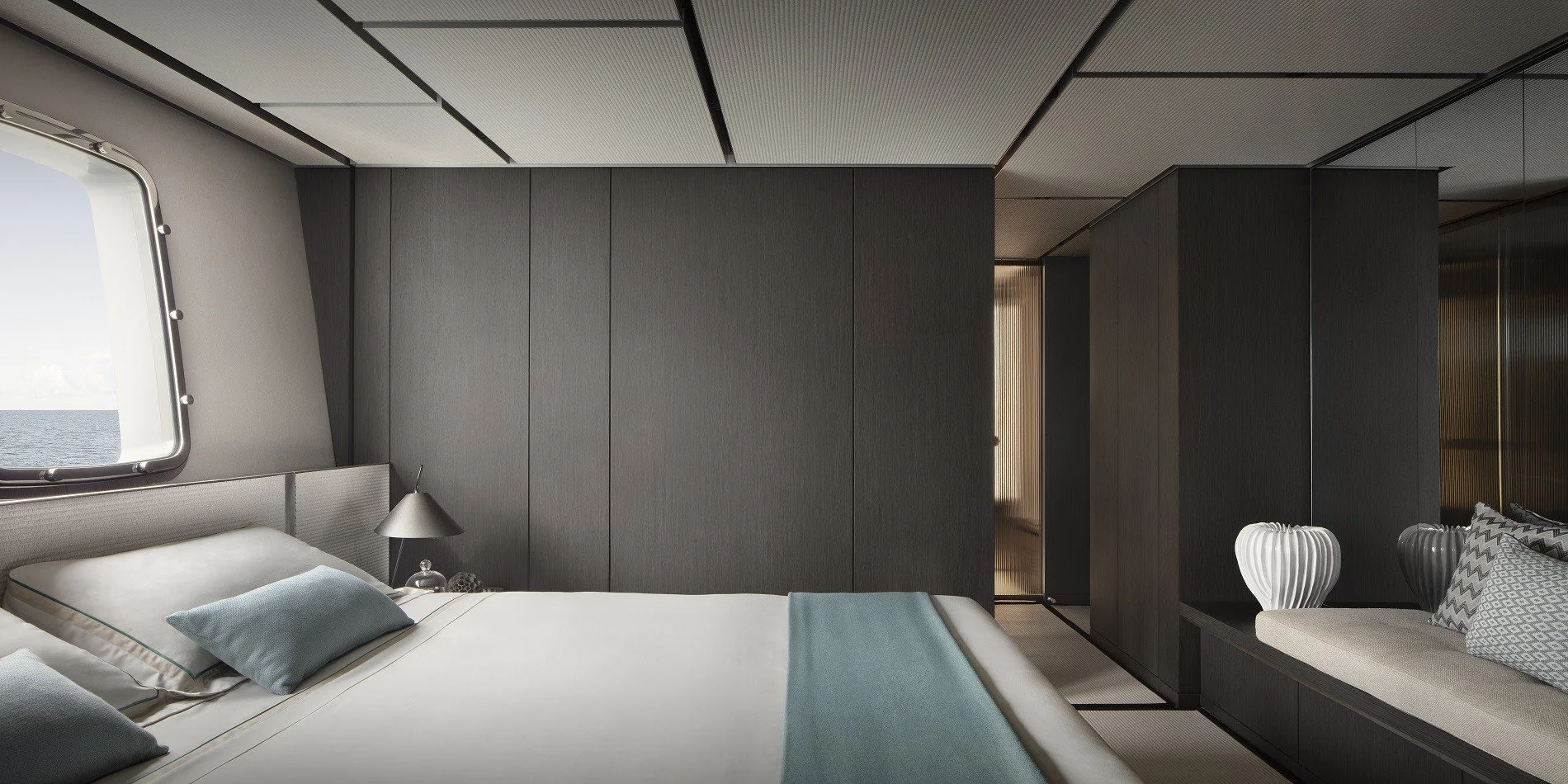 Modern bedroom with dark wood-paneled walls, a window showing the ocean, a neatly made bed with light blue pillows and blanket, a small bedside lamp, and a sitting area with decorative vases and pillows.