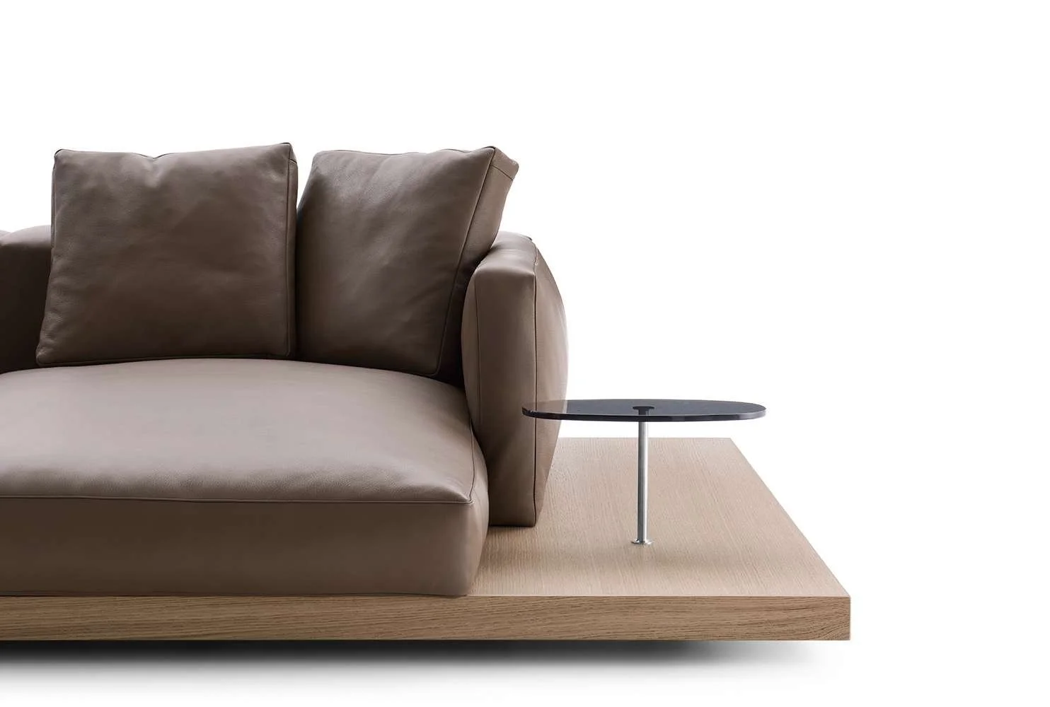 Close-up of a beige sofa with brown cushions on a wooden platform, with a small black side table attached to the armrest, against a plain white background.