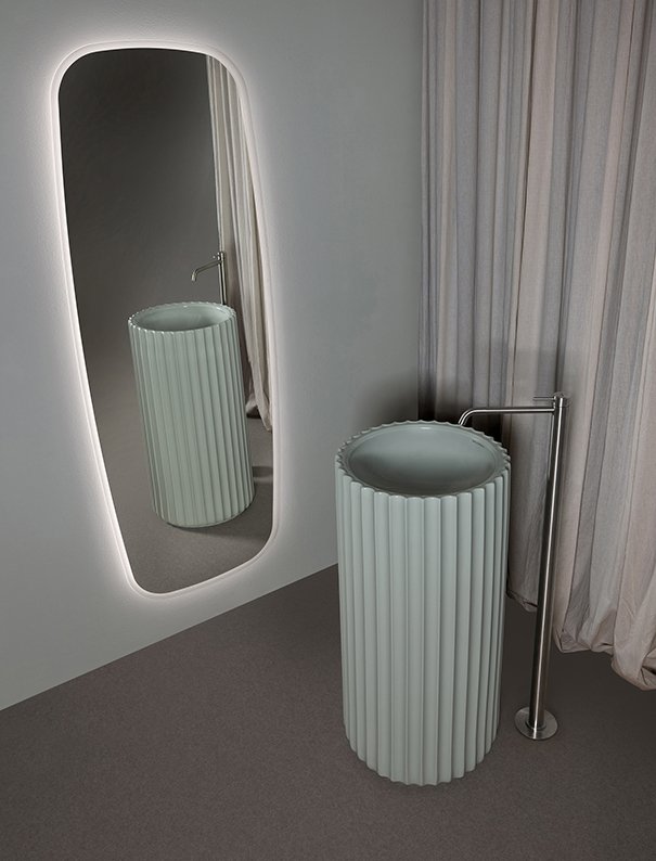 A white, cylindrical, ribbed sink with a silver faucet next to a large oval mirror with a backlit frame, set against light-colored walls and curtains.