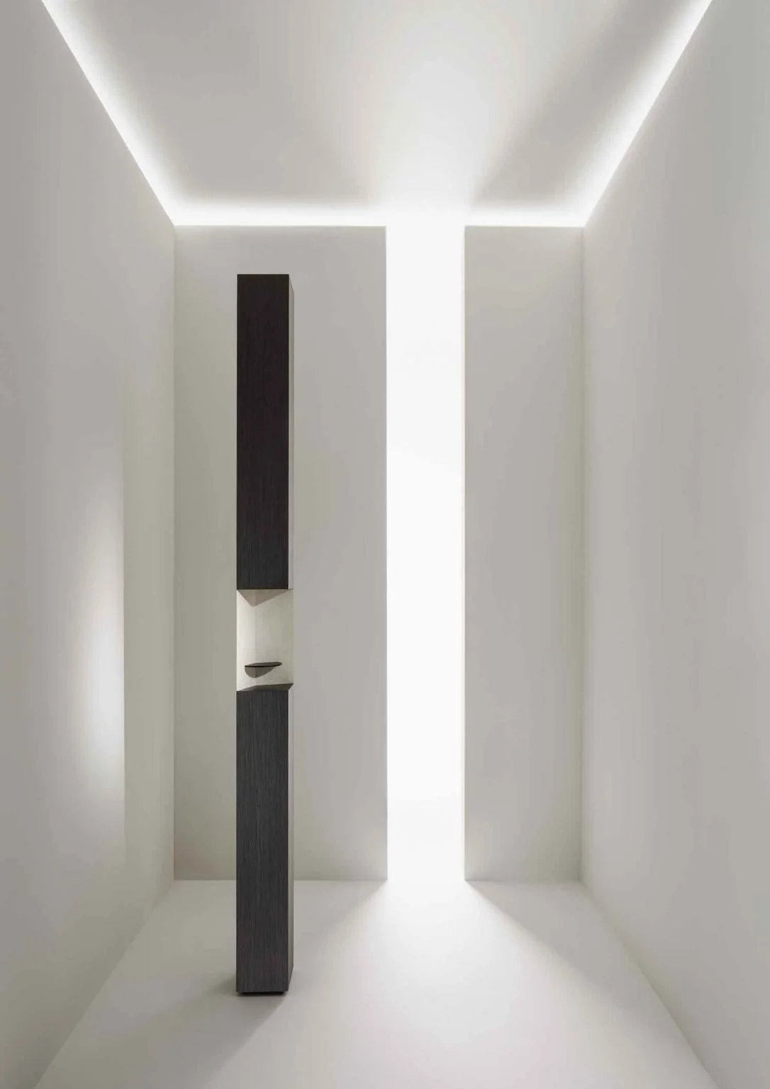 Minimalist interior with a tall dark vertical sculpture or panel and a small shelf in a white room with indirect ceiling lighting.