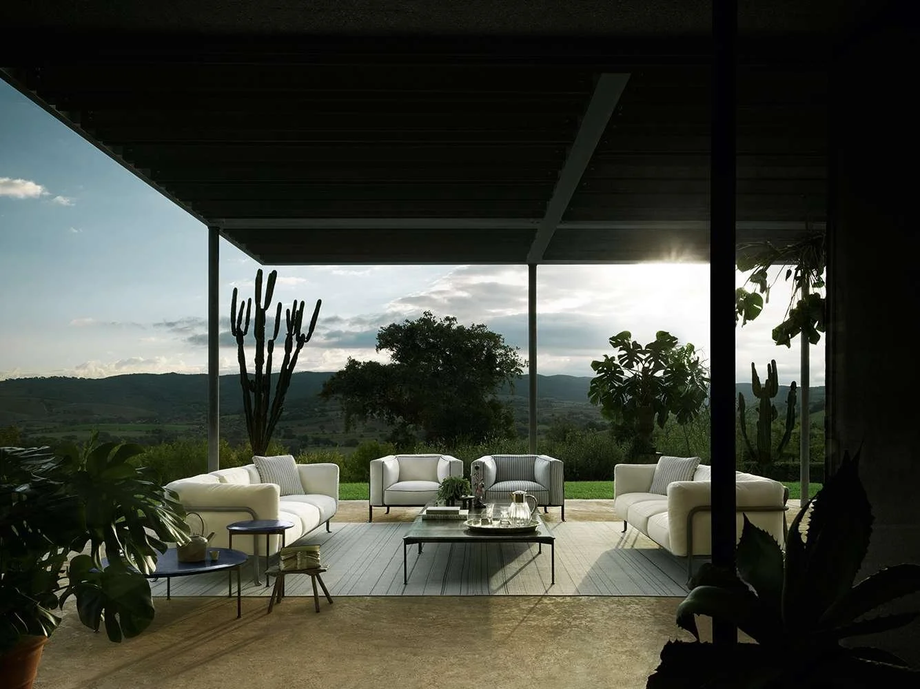 Outdoor patio with white sofas, a coffee table, plants, and a view of mountains and trees at sunset