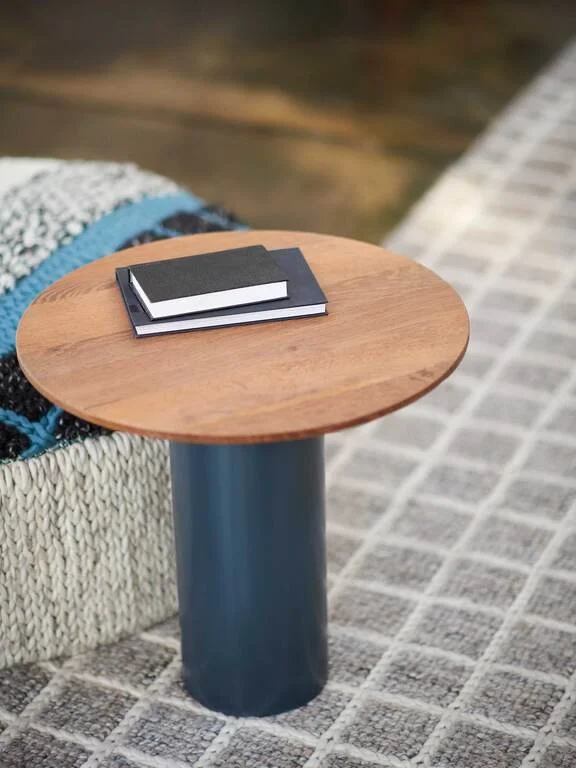 A small round wooden side table with a black and white notebook on top, placed next to a textured fabric sofa on a tiled floor.