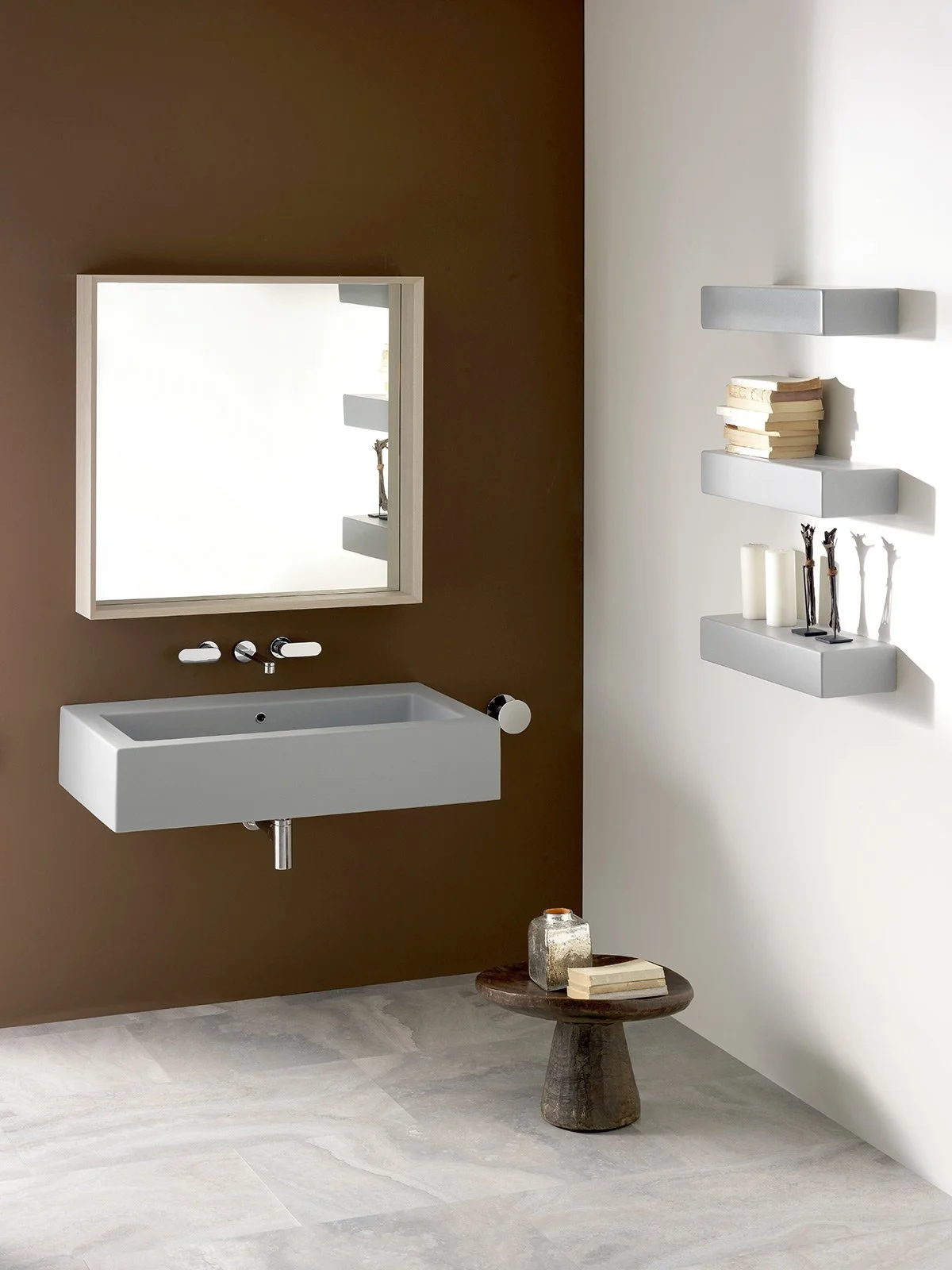 Minimalist bathroom with a white rectangular sink, mirror, and two white shelves holding books and decorative items, with a small round wooden side table and stone flooring.