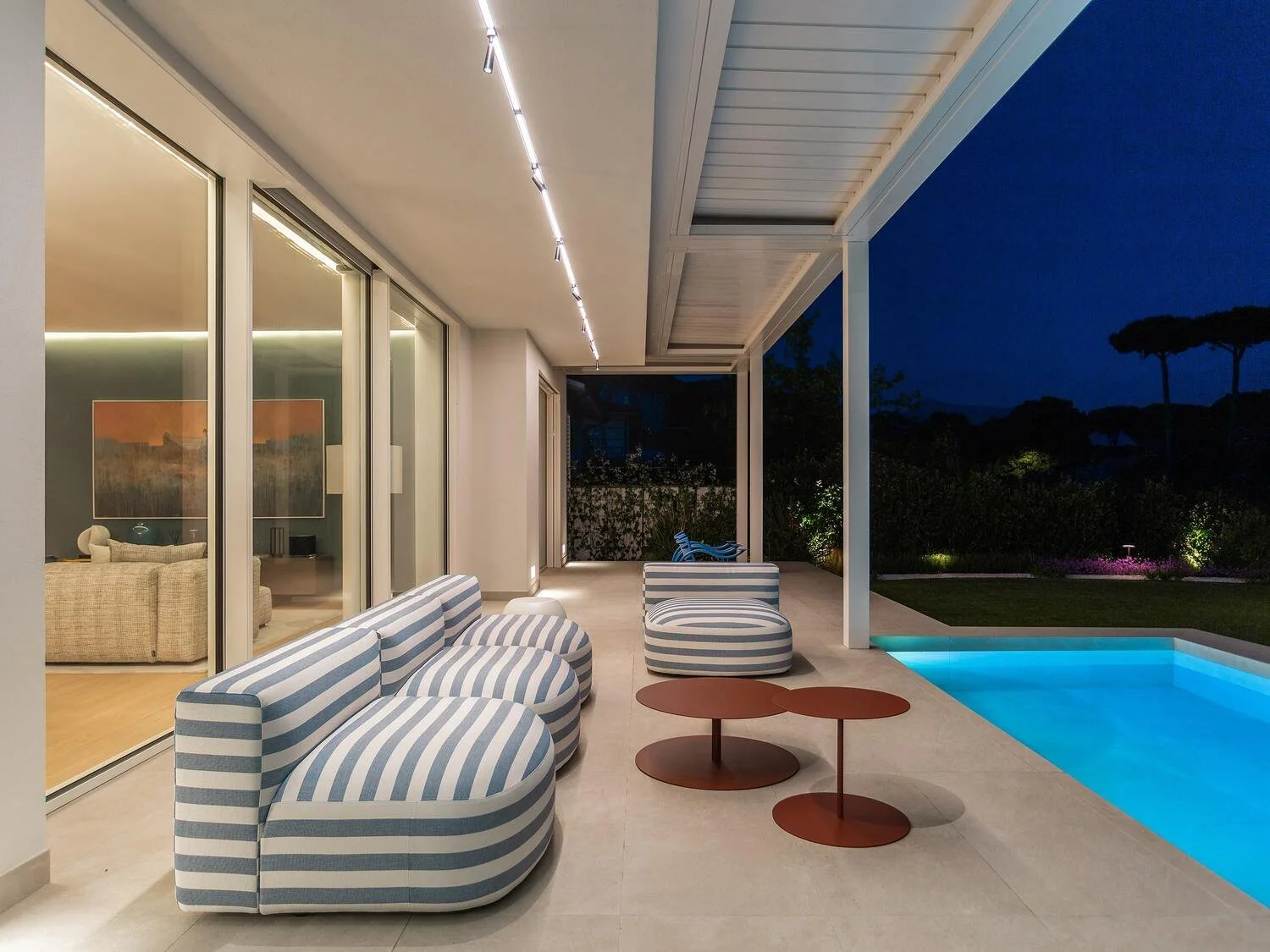 Modern patio with striped outdoor chairs, a small round side table, a swimming pool, and a view of trees and night sky.