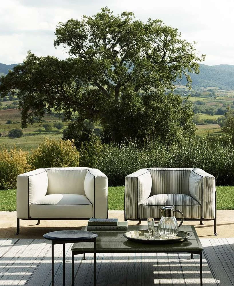 A patio or outdoor seating area with two modern armchairs, one plain white and one with striped fabric, placed outdoors against a backdrop of trees and rolling hills.