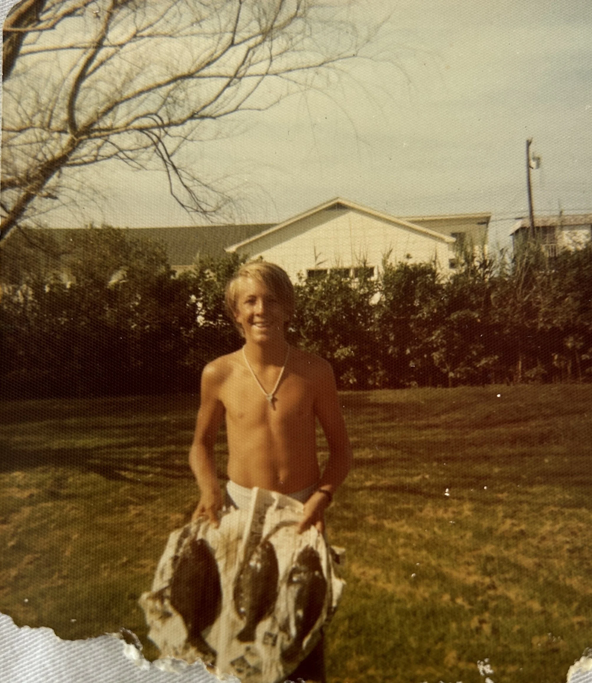 A shirtless young man with blonde hair smiling outdoors, standing in front of a hedge and a house with a pitched roof, during daytime.