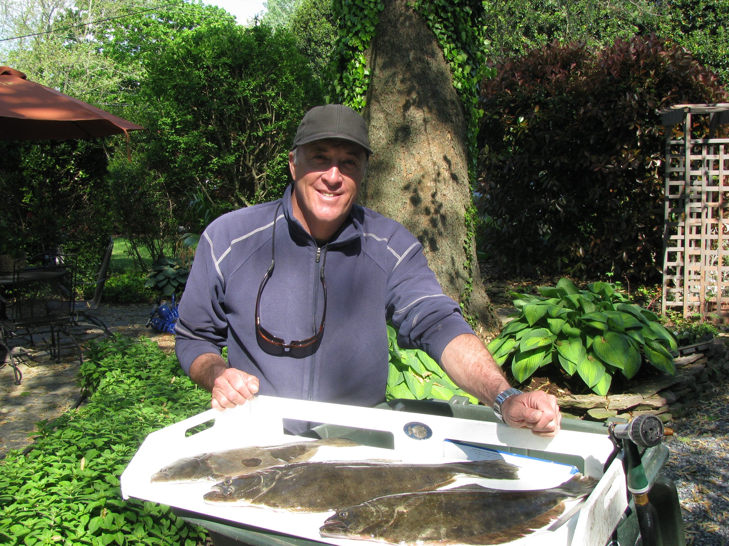 A smiling man wearing a gray jacket and black cap standing outdoors behind a white tray with two large fish on it, in a garden with lush green trees and plants. sandbar Captain Tilly OC Fishing Charters. Ocean City, MD