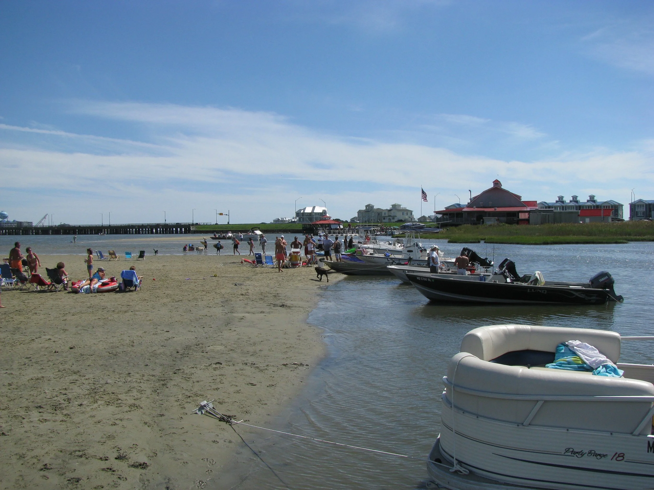 Beach scene with people relaxing on chairs, children playing, boats docked near the shore, a pier, and buildings in the background under a partly cloudy sky.  Nature tours, sightseeing. Captain Tilly OC Fishing Charters. Ocean City, MD