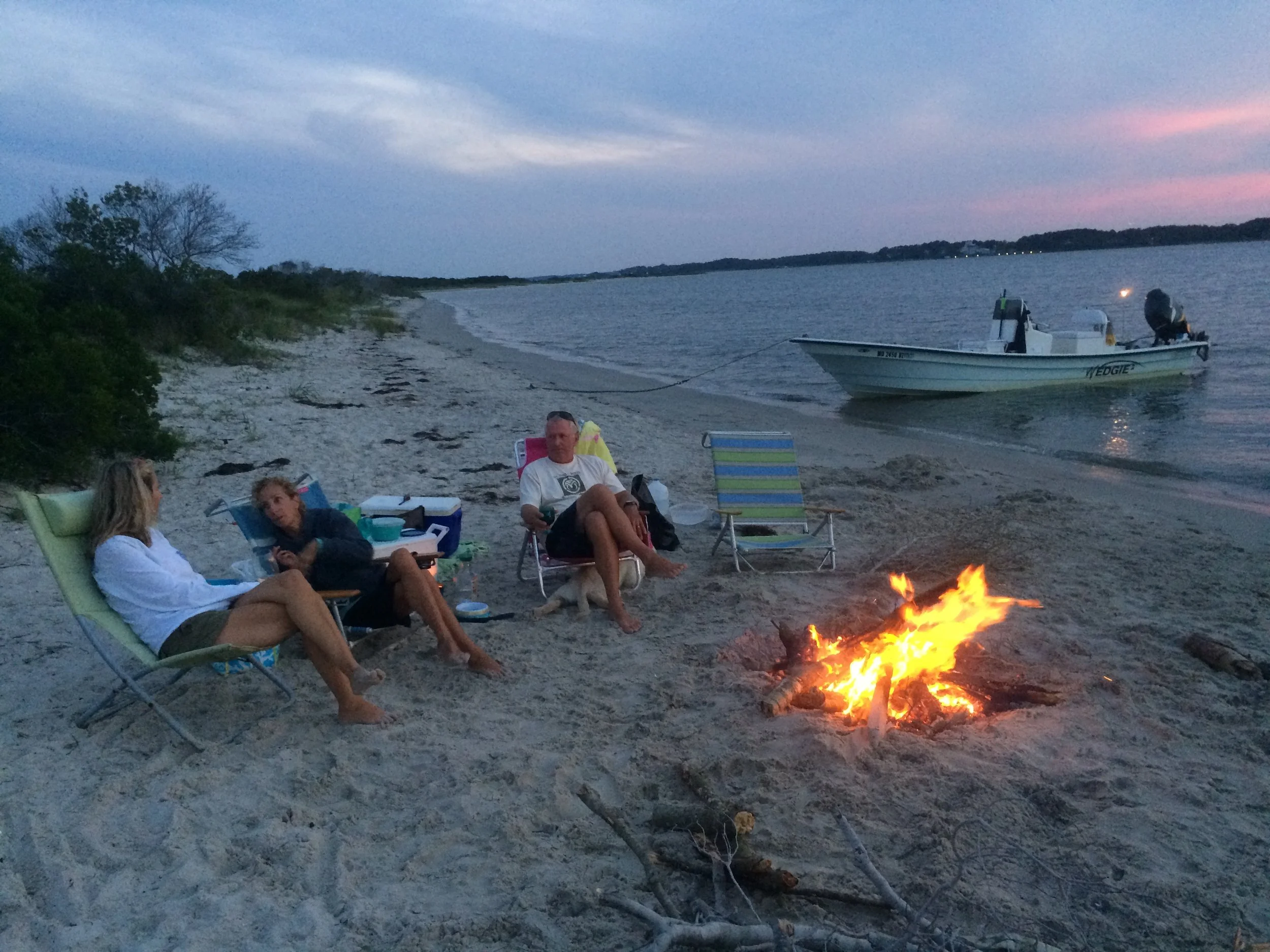 People relaxing on the beach around a campfire during dusk near a body of water, with a boat anchored nearby and a colorful sky in the background.  sandbar Captain Tilly OC Fishing Charters. Ocean City, MD