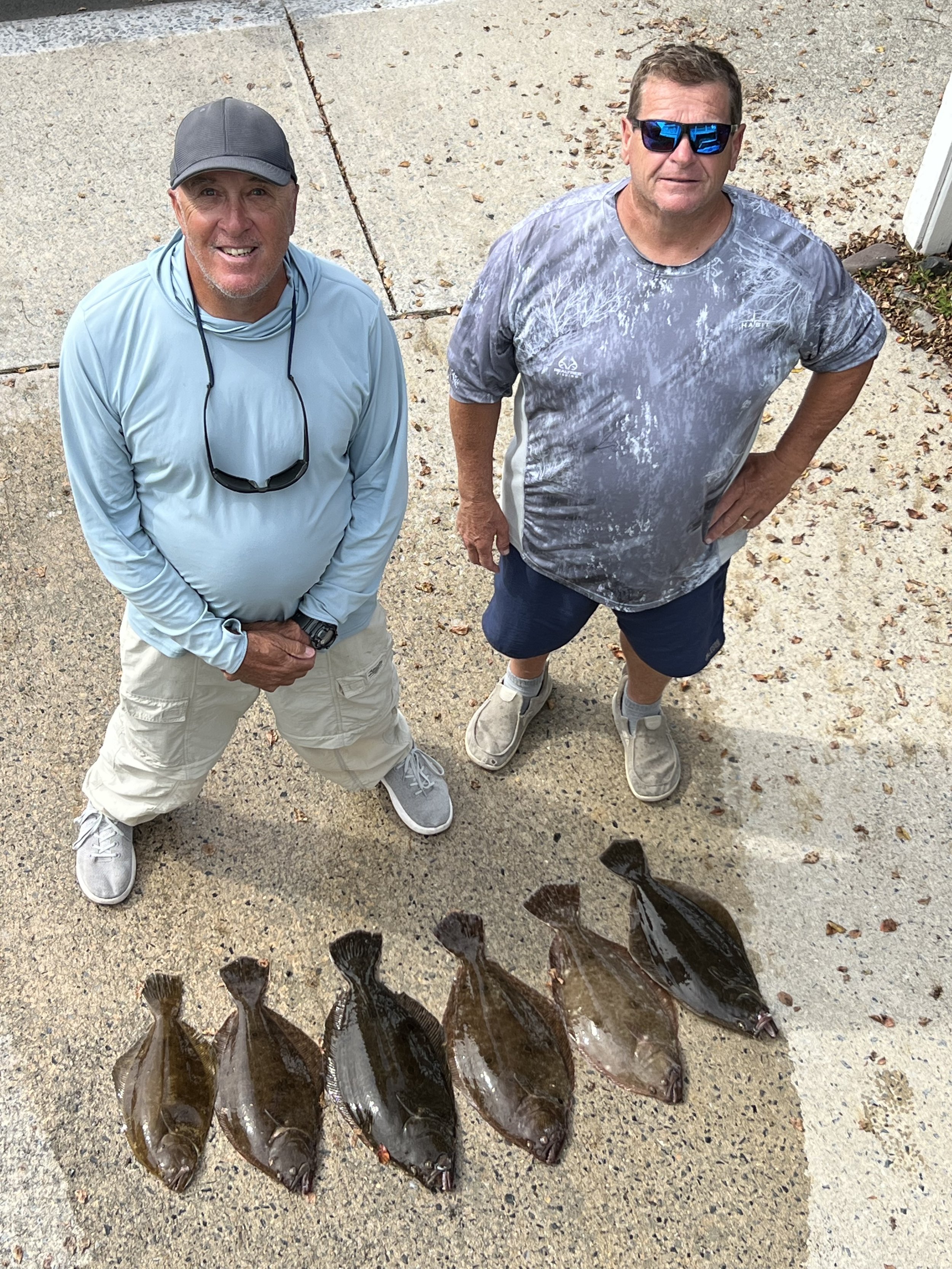Two men standing outdoors on a concrete surface, with four large flatfish on the ground in front of them, and a fifth fish tail visible at the bottom of the image. sandbar Captain Tilly OC Fishing Charters. Ocean City, MD
