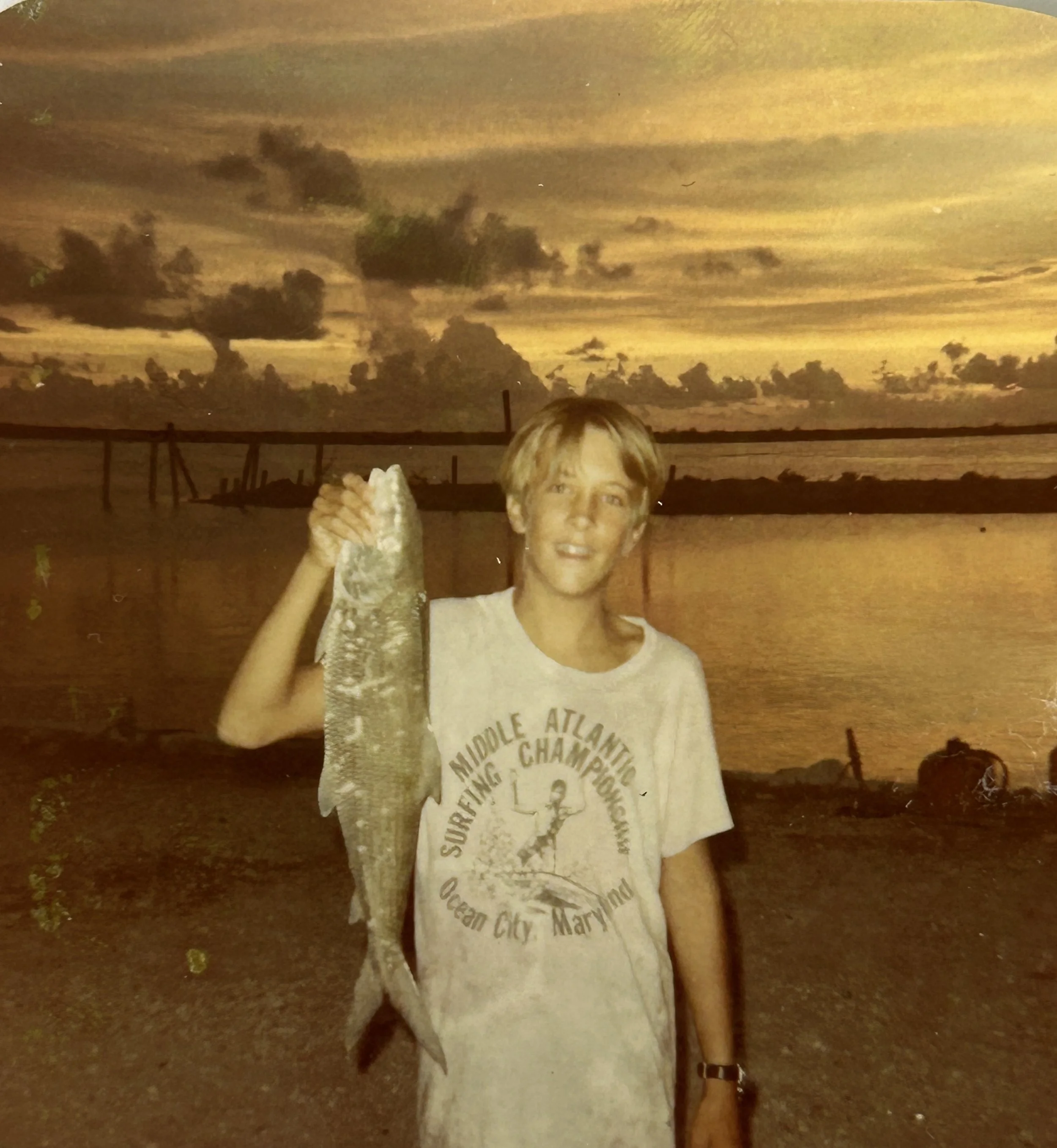A young boy standing outdoors at dusk, holding a large fish in his right hand and smiling at the camera. The background shows a wooden fence and a cloudy sky with a warm, yellowish hue.