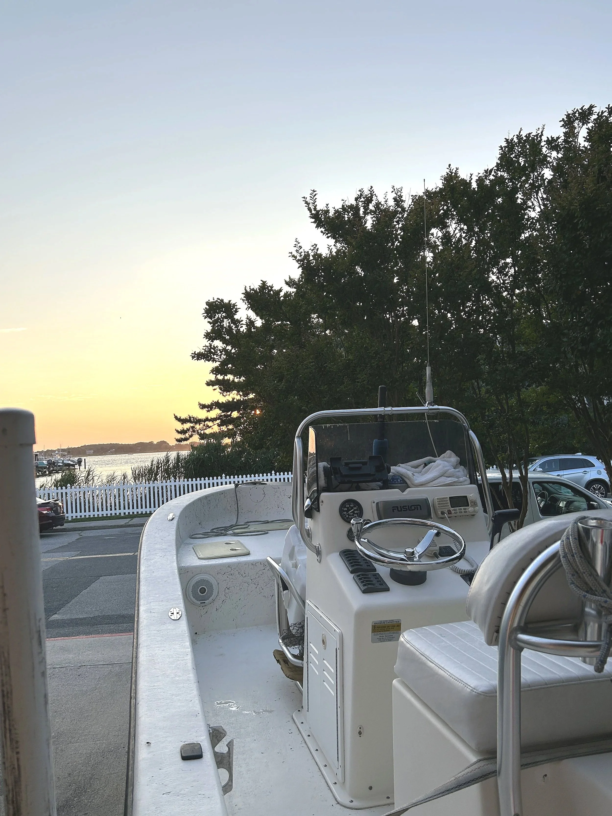 View of a boat dashboard with steering wheel and controls, parked outside near trees and a body of water, with a sunset sky.