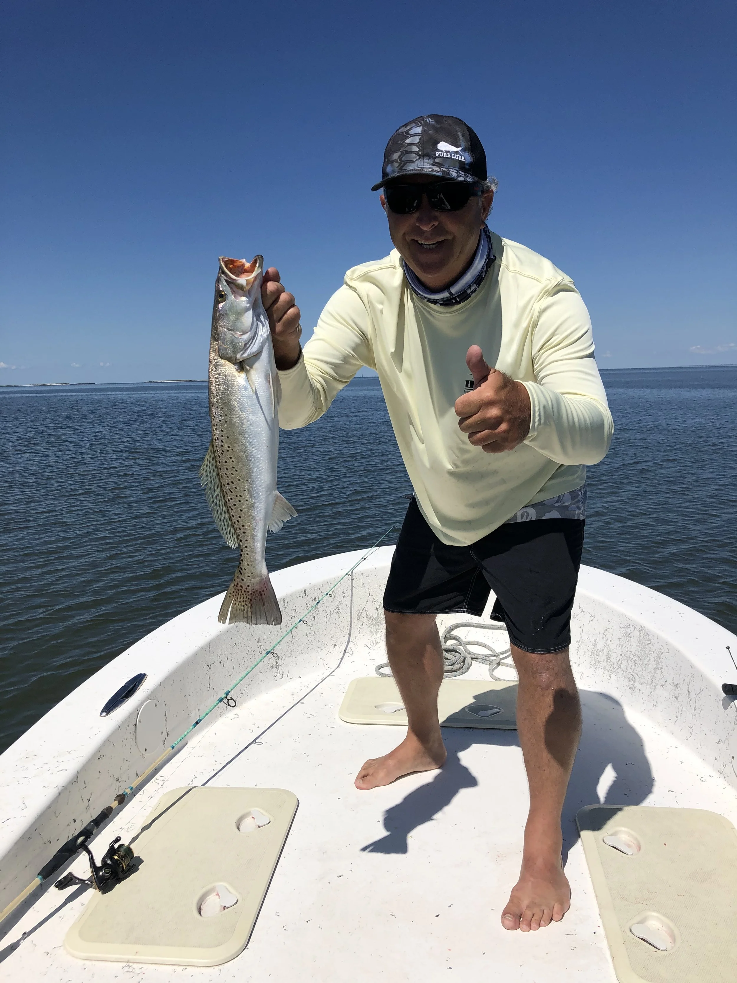 A man standing barefoot on a boat holding a large fish, with fishing rods lying on the boat's floor and a calm body of water in the background under a clear blue sky.