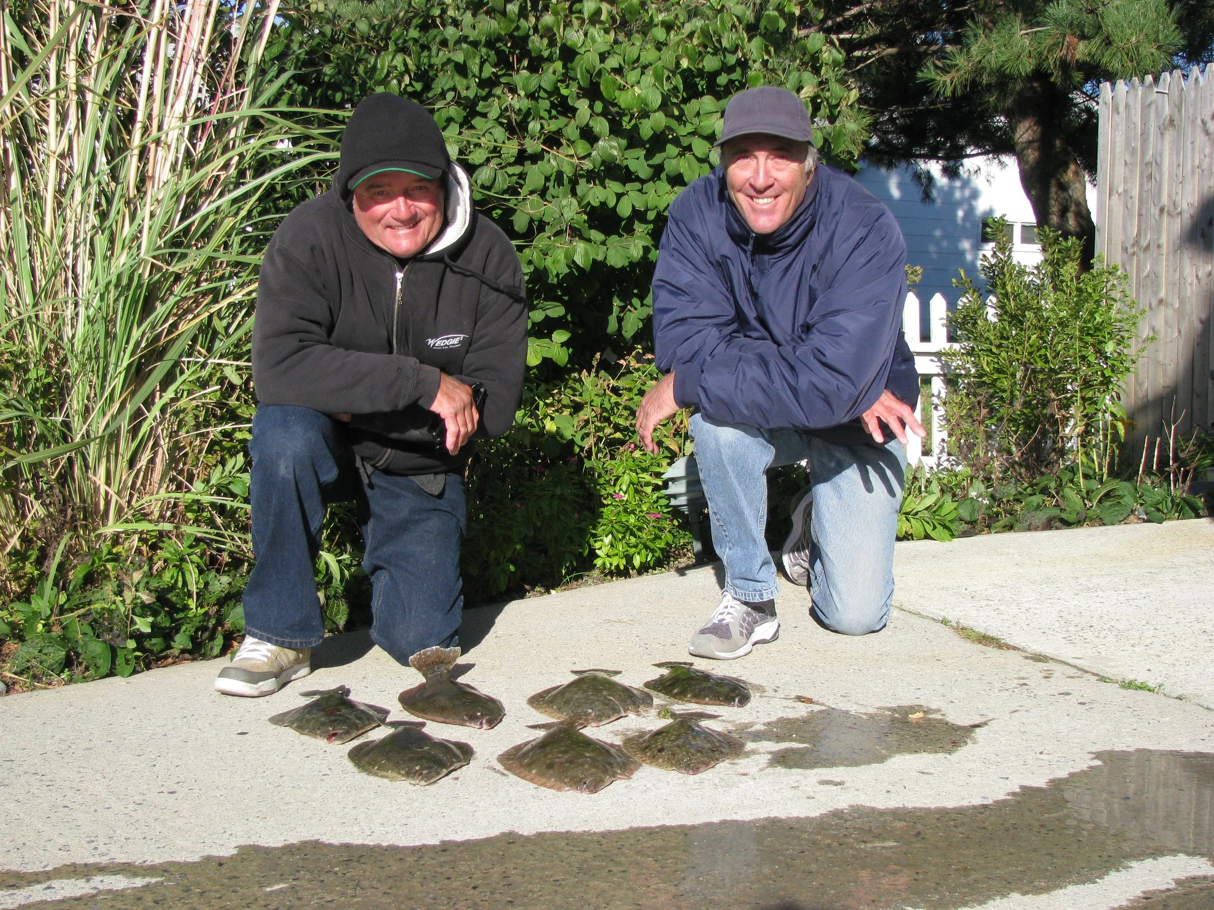 Two men crouching on a sidewalk outdoors, with a row of flat, dark-colored shells in front of them. sandbar Captain Tilly OC Fishing Charters. Ocean City, MD